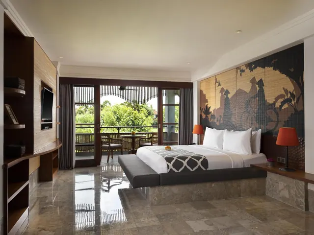 A bedroom with a king bed, a wall-mounted television, and glass doors opening onto a balcony with chairs and a view of trees.