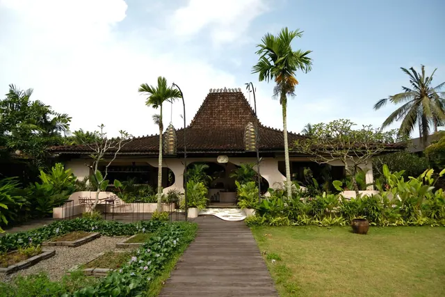 A traditional tiled-roof building stands at the end of a wooden walkway, flanked by palm trees and tropical planting.