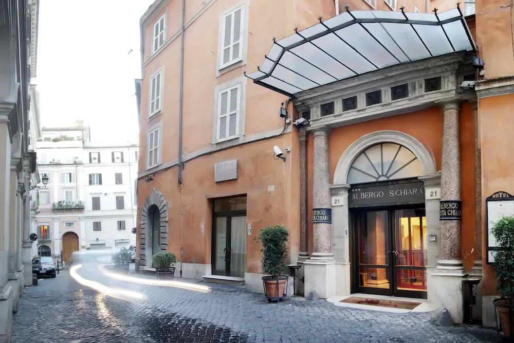 The hotel entrance under a glass canopy, with the sign Albergo Santa Chiara on the orange facade and a cobbled street in front.