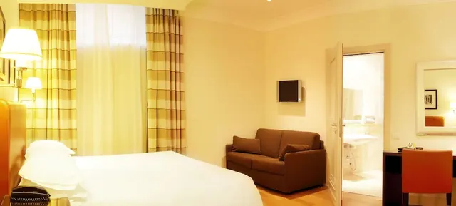 A bedroom with a large bed, a brown sofa, a wall-mounted television and a desk with an orange chair, with an open door to a bathroom sink.
