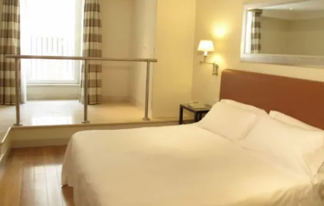 A bedroom with a made double bed with white linens and a brown headboard, a wall lamp and mirror, and a raised area with a metal railing beside a window with checked curtains.