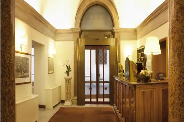 A hotel lobby with a brass-framed glass lift, a wooden reception counter with a table lamp and flowers, and a potted orchid on a marble pedestal.