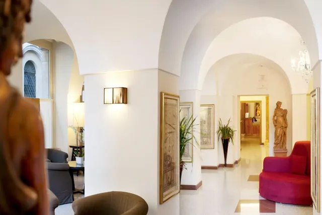 A hotel lobby corridor with vaulted arches, framed prints, a statue, a red armchair and potted plants.
