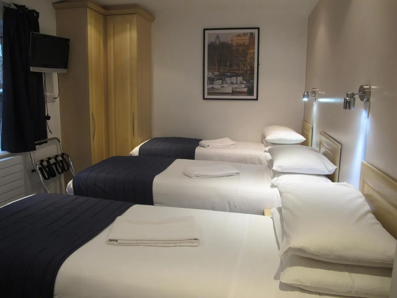 A bedroom with three single beds with navy throws and folded white towels, a wooden wardrobe, a wall-mounted television and a framed print of a London river scene.