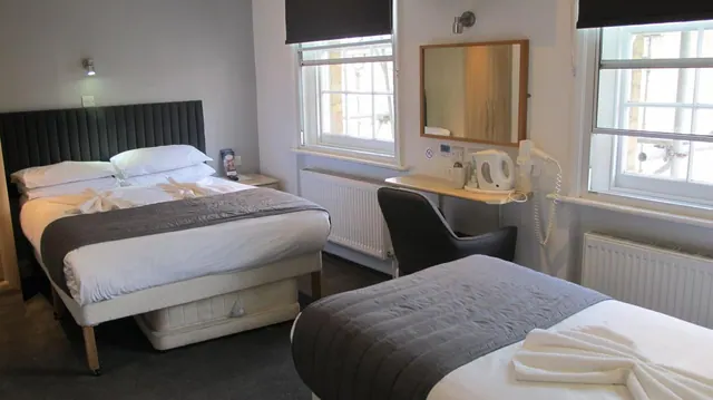 A hotel bedroom with a double bed, a single bed, a desk with a kettle and mirror, and windows facing a neighbouring building.