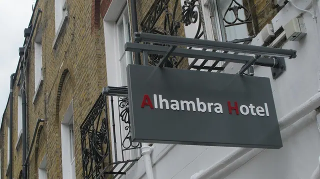 A grey hanging sign reading Alhambra Hotel is mounted on a white facade beside a yellow brick building with wrought iron railings under an overcast sky.