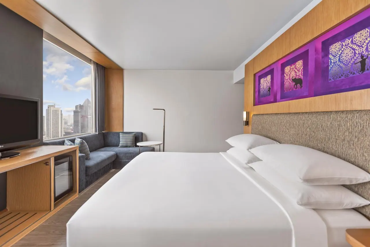 A hotel bedroom with a made king bed, a grey sofa by the window, a wall-mounted television and a small fridge, with high-rise buildings visible outside.