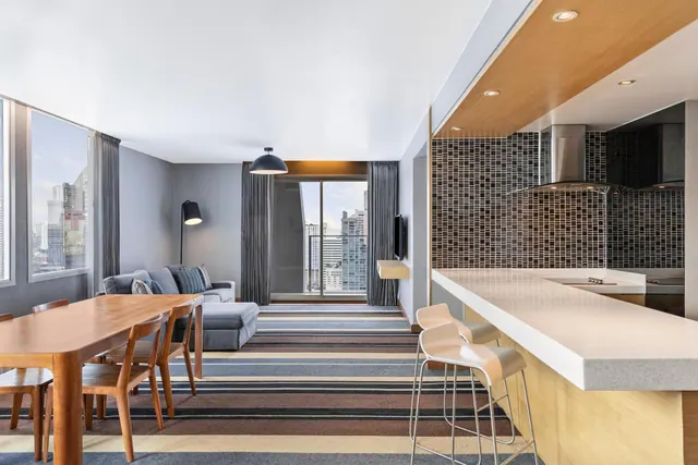 A living and dining area with a grey sofa, a wooden dining table, bar stools at a kitchen counter, and high-rise buildings visible through the windows.