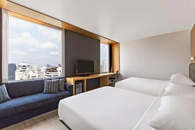 A hotel room with two white beds, a blue sofa, a television on a wooden desk, and a large window showing city high-rise buildings.