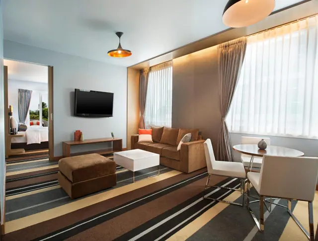 A modern living area with a brown sofa, ottoman, white coffee table, wall-mounted television and white dining chairs, with a bedroom visible through the sliding door.