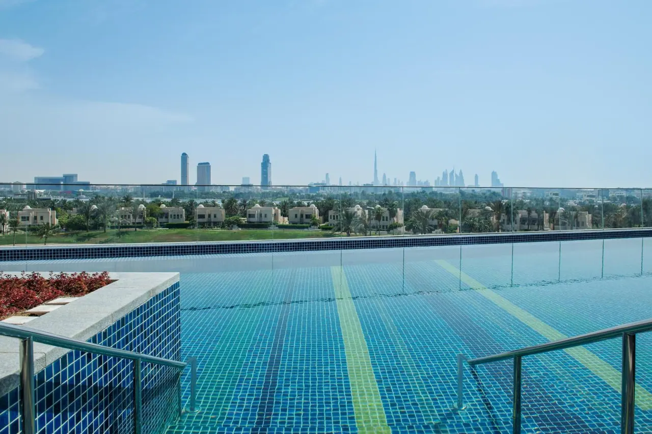A rooftop swimming pool with a glass railing overlooks low-rise villas, palm trees and the Dubai skyline, including the Burj Khalifa, under a hazy blue sky.