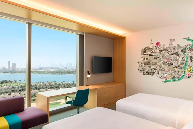 A twin-bedded hotel room with floor-to-ceiling windows looking over a lake, trees and a city skyline with the Burj Khalifa in the distance.