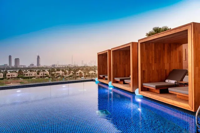 A rooftop pool with an edge overlooking a hazy city skyline, with wooden cabanas containing sun loungers along the right side.