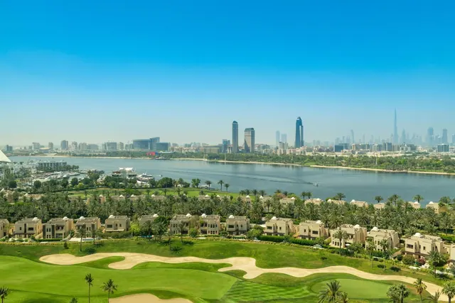 Aerial view over a golf course and low-rise villas toward a waterway and a city skyline with a tall spire visible in the distance.