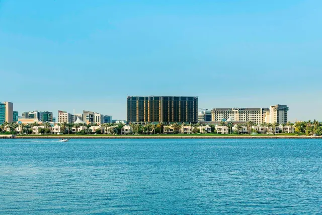 Low white villas, palm trees and several tall hotel and office buildings line the far shore of a calm blue waterway under a clear sky, with a small boat on the water.