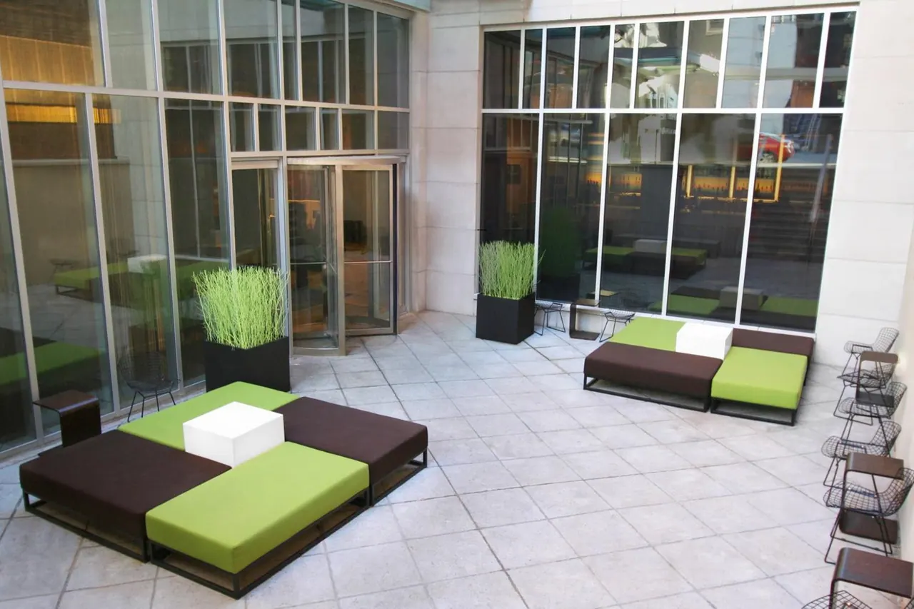 A paved outdoor courtyard with green and brown modular lounge seating, white cube tables, planters with tall grasses, and glass walls.