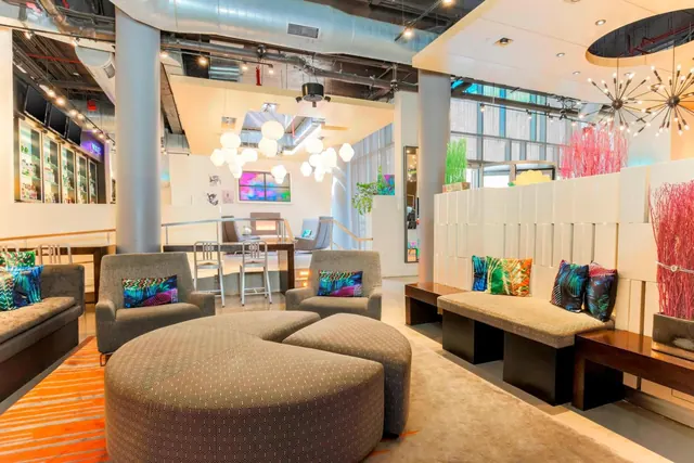 A lounge area with grey upholstered ottomans, armchairs and benches, colourful patterned cushions, and globe pendant lights, with a bar along the left wall.