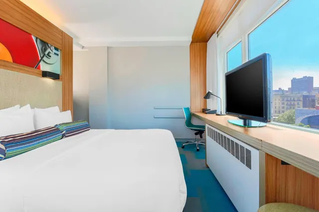 A hotel bedroom with a king bed, a flat-screen TV and a desk beside a window overlooking city buildings under a blue sky.