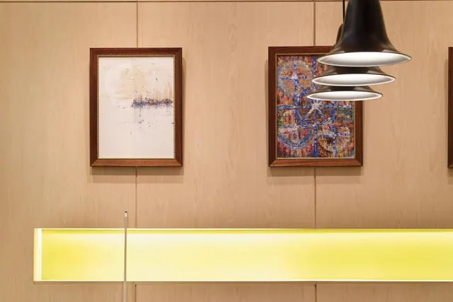 Two framed abstract paintings hang on a wood-paneled wall above a glowing yellow ledge, with black pendant lamps hanging on the right.