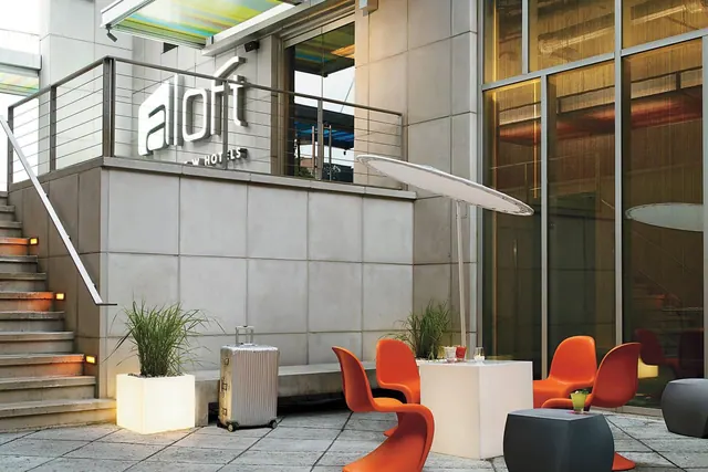 An outdoor terrace with orange chairs, white cube tables, a white umbrella, potted plants and a staircase beside an Aloft hotel entrance, with a suitcase on the ground.