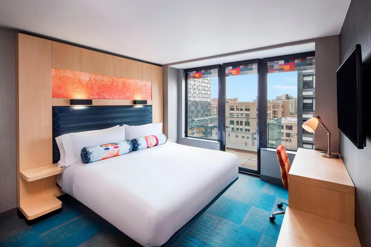 A bedroom with a king bed, a wall-mounted television, a work desk and floor-to-ceiling windows overlooking city buildings.