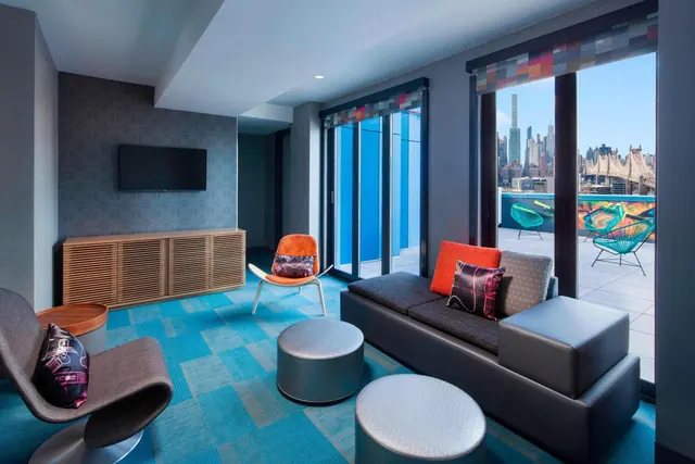 A lounge area with a sofa, ottomans, a wall-mounted television and a wood sideboard, with sliding glass doors opening onto a terrace and a view of the Manhattan skyline and the Queensboro Bridge.