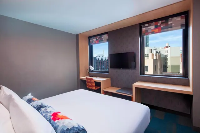 A bedroom with a made bed, a desk with an orange chair, a wall-mounted television and two windows looking out onto city buildings.