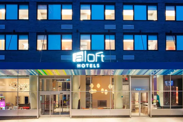 Exterior of an Aloft Hotels building at dusk, with illuminated windows, a striped entrance awning and glass doors at street level.