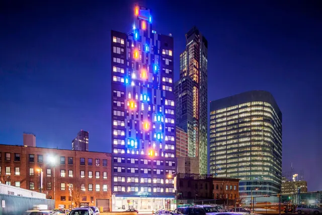 Night exterior of a tall tower with colored lights on its facade and an Aloft sign at street level, with taller buildings and a lit office block behind it.
