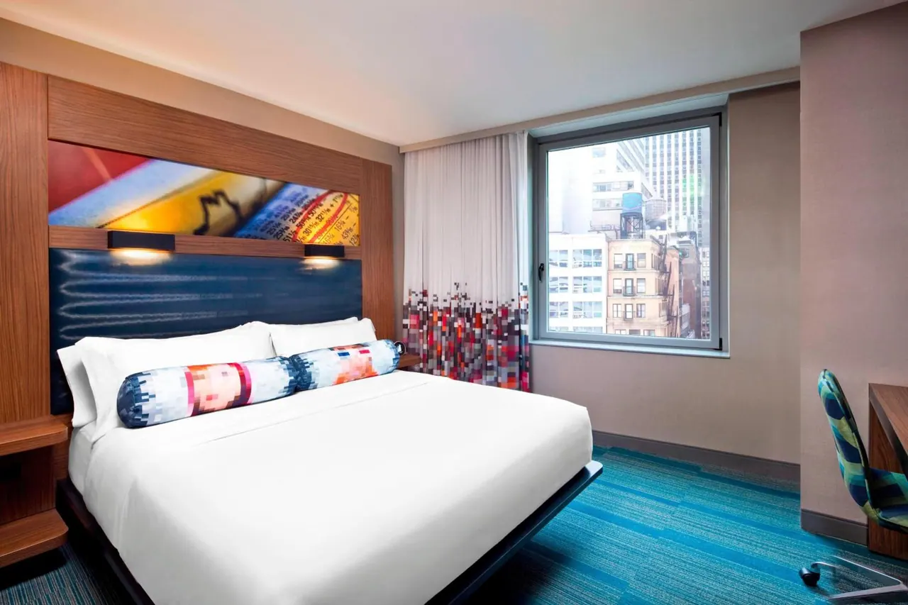 A bedroom with a king bed under a colourful headboard, a window looking out over city buildings and a rooftop water tank, and a desk with a chair on the right.