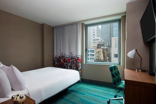 A hotel bedroom with a made bed, a wall-mounted television, a desk with a lamp and a green chair, and a window showing city buildings.