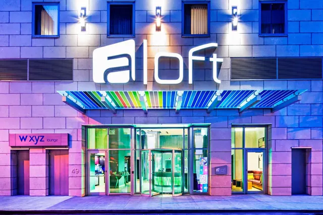 Exterior of the Aloft hotel entrance at dusk, with an illuminated sign, a striped multicolour canopy, revolving doors and a W XYZ lounge sign beside the entrance.