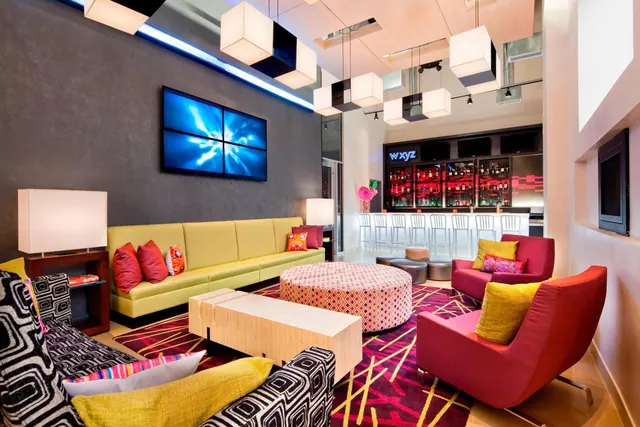 A lounge with a green sofa, red armchairs and a patterned rug, with a wall-mounted video screen and a bar with stools behind.
