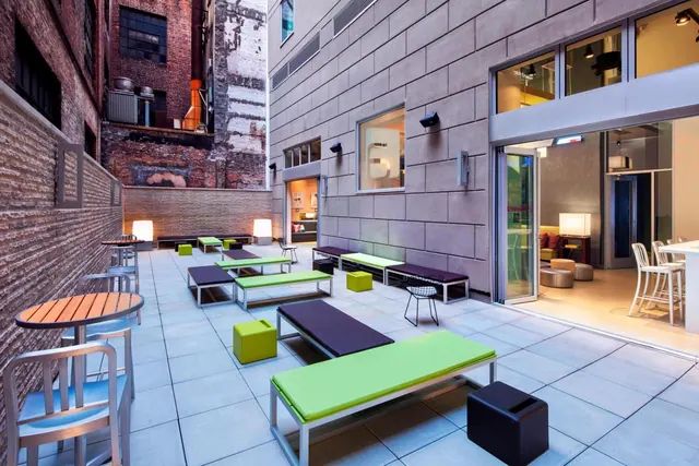 An outdoor courtyard terrace with green and purple lounge benches, round wooden tables and chairs, enclosed by brick and concrete walls, with an interior lounge visible through glass doors.