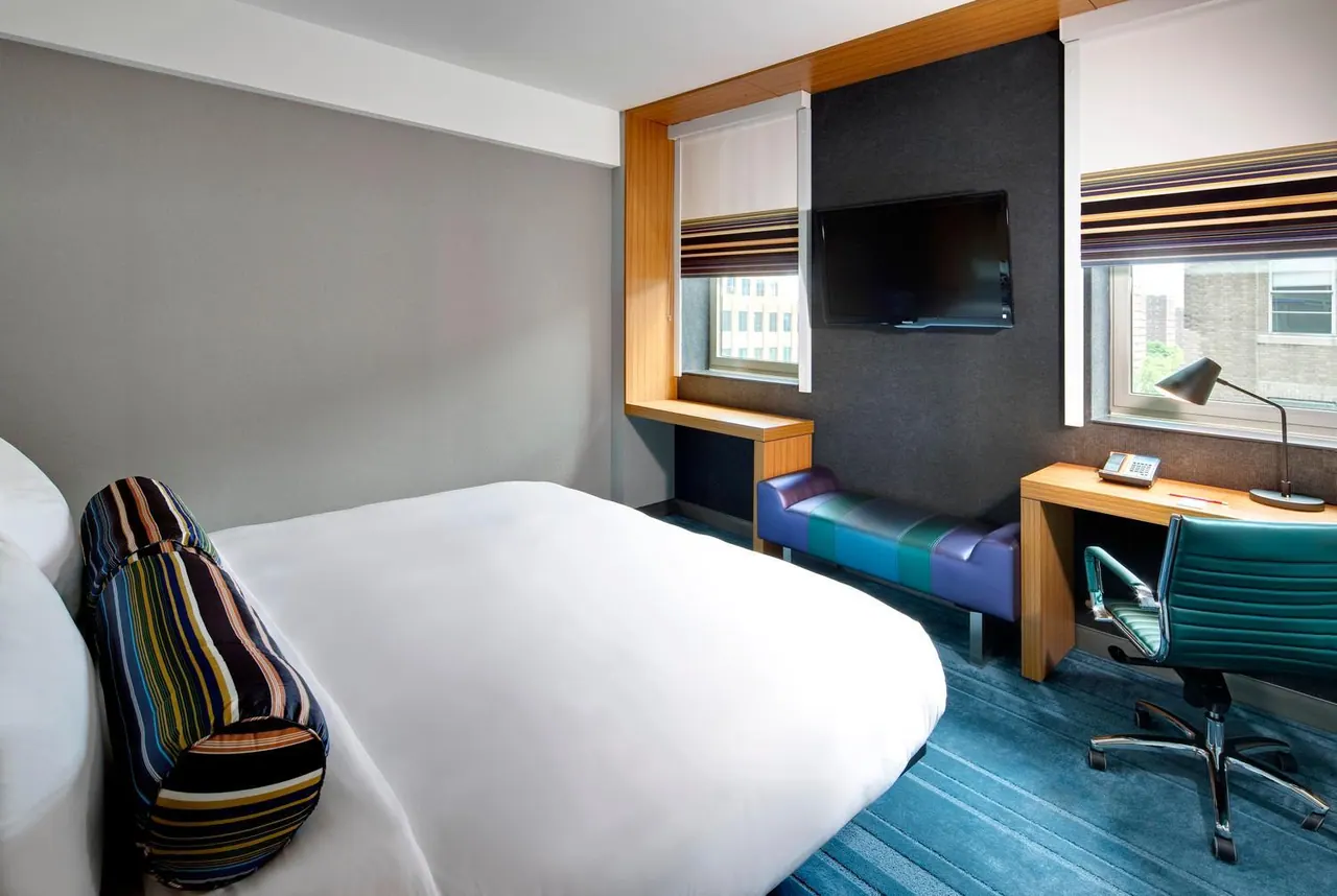 A hotel bedroom with a made double bed, a wall-mounted television, a desk with a chair and lamp, and windows looking onto nearby buildings.