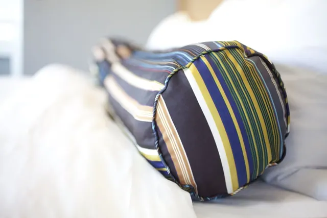 A striped bolster pillow lies on a bed with white linens in front of a blurred grey wall.