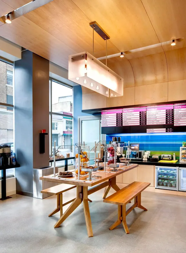 A breakfast area with a wooden communal table and benches, a counter with illuminated menu boards and a beverage fridge, and tall windows facing the street.