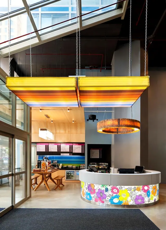 A lobby with a curved reception desk covered in a floral pattern, a striped yellow and orange canopy hanging overhead, and a café counter with menu boards behind, next to glass entrance doors.