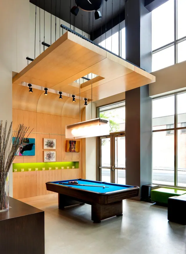 A lobby lounge with a blue felt pool table beneath a suspended wood ceiling feature, green and black seating, and tall windows facing a street.