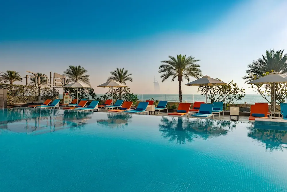 Outdoor pool with orange and blue sun loungers and parasol umbrellas, palm trees, and the sea with the Burj Al Arab visible in the distance.