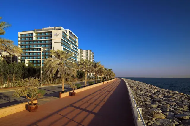 A wide paved promenade runs past palm trees and a white aLoft hotel building, with a rocky shoreline and the sea on the right.