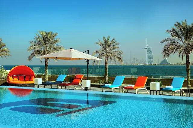 An outdoor pool with coloured loungers, a canopy daybed and a cantilever umbrella, with the sea and the Burj Al Arab visible in the distance.