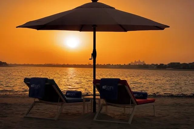 Two loungers with blue towels sit under a shade umbrella on a sandy beach facing the water at sunset, with a distant resort building silhouetted on the shoreline.