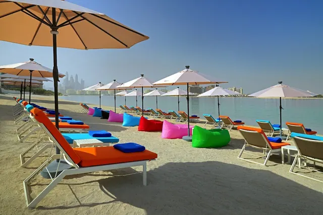 Rows of orange and white loungers, colourful bean bags and parasol umbrellas stand on a sandy beach beside calm turquoise water, with a city skyline in the distance.