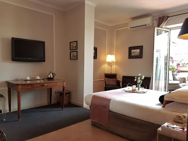 A hotel bedroom with a double bed dressed in white linen, a wall-mounted television, a wooden desk with a kettle, and open glass doors onto a balcony with flowers and chairs.