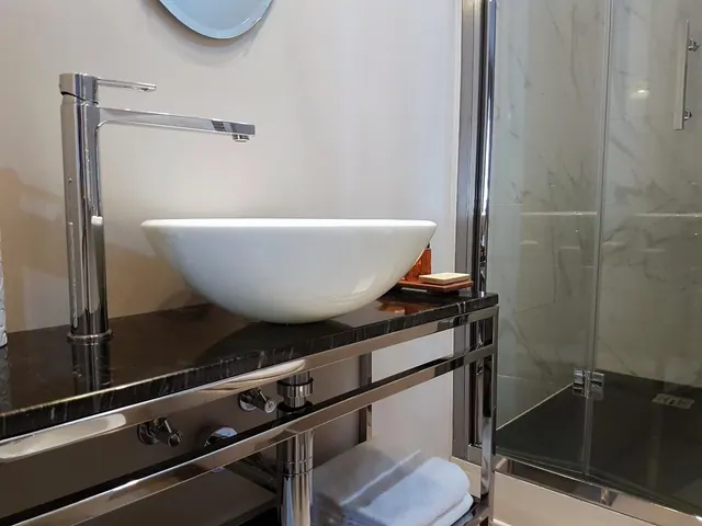 A white vessel sink sits on a black marble vanity beside a tall chrome tap, with a glass-walled shower and folded towels on the lower shelf.