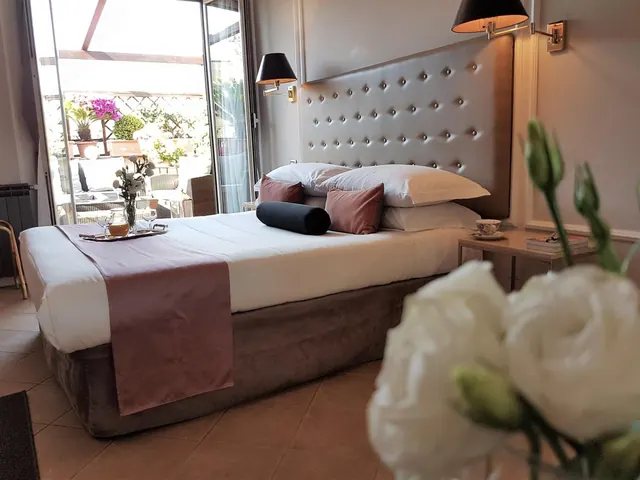 A made double bed with a tufted headboard and a serving tray, with glass doors opening onto a terrace with potted plants and wicker chairs.