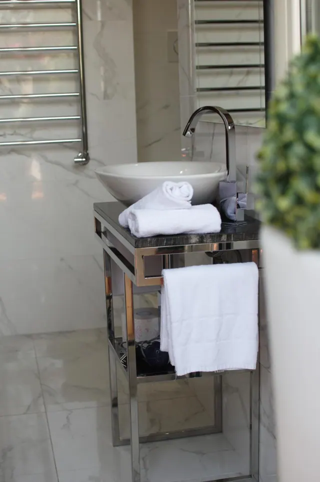 A bathroom with a white vessel sink on a chrome stand, folded white towels, a heated towel rail and marble-effect wall tiles.