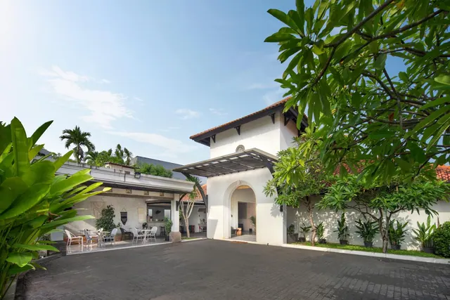 A white building with an arched entrance and a covered canopy stands beside a paved driveway, with frangipani trees and a terrace with chairs visible on the left.