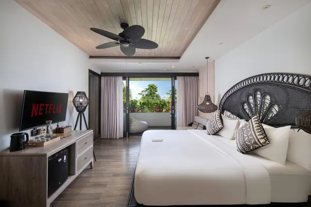 A bedroom with a king bed, a wall-mounted television, a ceiling fan, and glass doors opening onto a balcony with a hanging chair and a view of green garden trees.